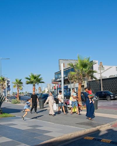 A sunny outdoor street scene showing a paved promenade with pedestrians, palm trees, a McDonald's sign, and modern buildings along a coastal road.