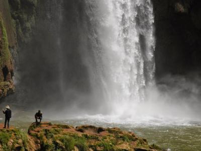 A large waterfall cascading into a pool with two people standing on rocks in the foreground.