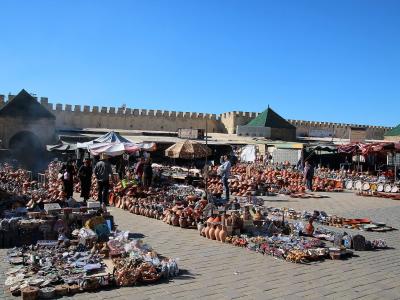 A sunlit public square paved with stone, filled with market stalls selling pottery and souvenirs, backed by a high crenellated wall with an arched gateway and green-roofed structures.