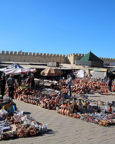 A sunlit public square paved with stone, filled with market stalls selling pottery and souvenirs, backed by a high crenellated wall with an arched gateway and green-roofed structures.