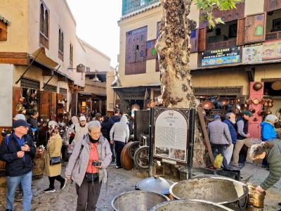 A sunlit stone square in a Moroccan medina filled with tourists and copper artisans, featuring large metal basins in the foreground and a prominent information sign mounted on a tree trunk.