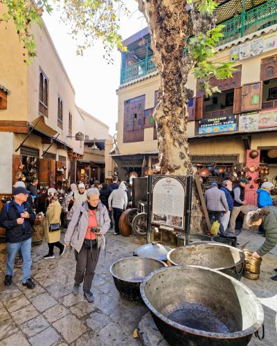 A sunlit stone square in a Moroccan medina filled with tourists and copper artisans, featuring large metal basins in the foreground and a prominent information sign mounted on a tree trunk.