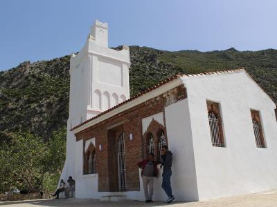 A white building with exposed brickwork and a tall, unfinished tower stands on a hillside with a rocky mountain in the background, featuring people standing and sitting near the entrance.