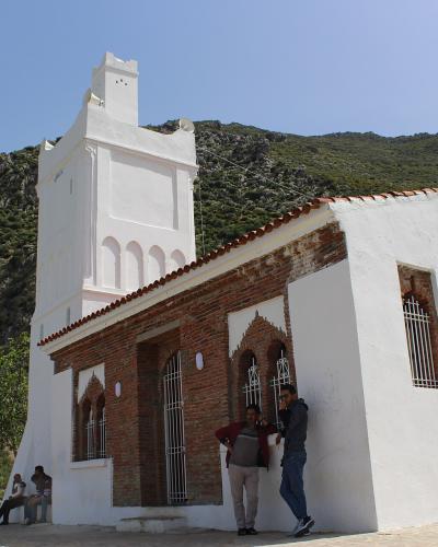 A white building with exposed brickwork and a tall, unfinished tower stands on a hillside with a rocky mountain in the background, featuring people standing and sitting near the entrance.