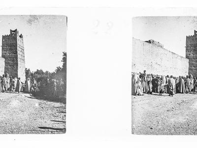 A vintage black-and-white stereoscopic photograph showing a group of men in traditional robes standing on a dirt path in front of a tall, fortified earthen wall and tower.