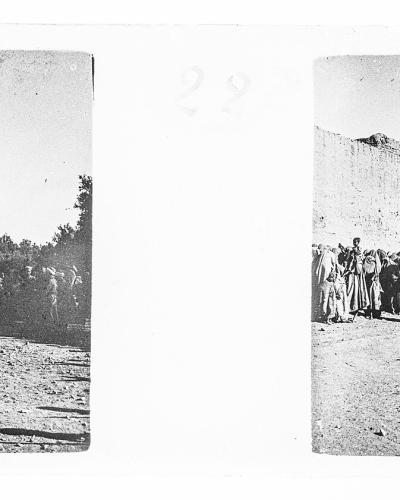 A vintage black-and-white stereoscopic photograph showing a group of men in traditional robes standing on a dirt path in front of a tall, fortified earthen wall and tower.