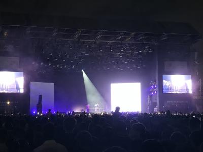 A night-time concert scene showing a large stage with multiple screens, lighting rigs, and a crowd of spectators, with the text "WWW.MAWAZINE.RA" visible on the lower sections of the stage structure.