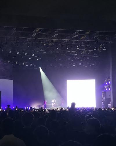 A night-time concert scene showing a large stage with multiple screens, lighting rigs, and a crowd of spectators, with the text "WWW.MAWAZINE.RA" visible on the lower sections of the stage structure.