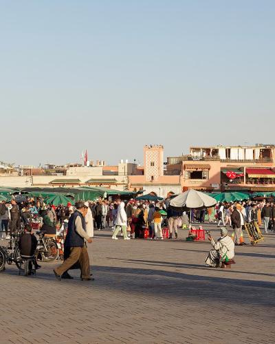 A wide-angle view of a large, paved public square crowded with people, green market umbrellas, and surrounding terracotta-colored buildings under a clear sky.