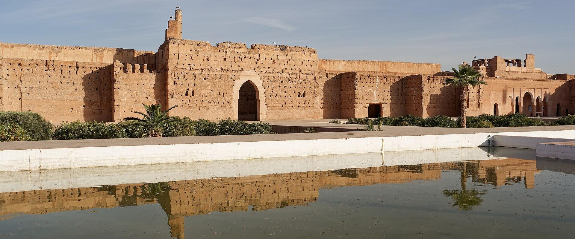 A wide shot of the ruins of a large, ochre-colored palace complex with crenellated walls, arched gateways, and a long reflecting pool in the foreground under a blue sky.