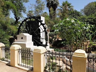 A large black noria (water wheel) set within a white stone structure, surrounded by lush green trees and banana plants, viewed from behind a cream-colored fence with black iron railings.