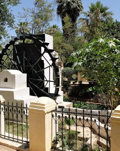 A large black noria (water wheel) set within a white stone structure, surrounded by lush green trees and banana plants, viewed from behind a cream-colored fence with black iron railings.