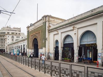 A street-level view of a long, white building with a series of arches, green tiled accents, and a tram line running in the foreground, located in Casablanca.