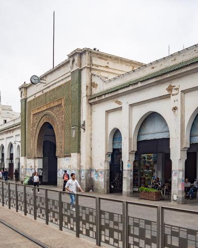 A street-level view of a long, white building with a series of arches, green tiled accents, and a tram line running in the foreground, located in Casablanca.
