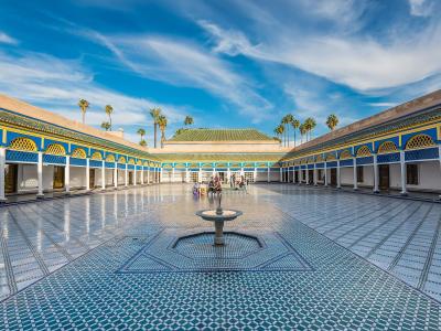 A wide-angle view of a large, rectangular courtyard with intricate blue and white zellige tilework on the floor, surrounded by arcades with white columns and blue and yellow decorative arches, featuring a central fountain and palm trees in the backgr