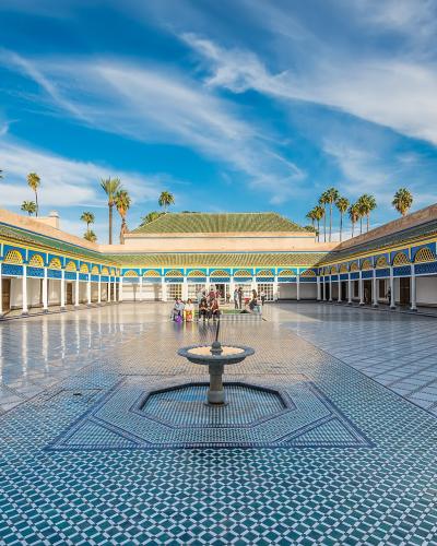 A wide-angle view of a large, rectangular courtyard with intricate blue and white zellige tilework on the floor, surrounded by arcades with white columns and blue and yellow decorative arches, featuring a central fountain and palm trees in the backgr