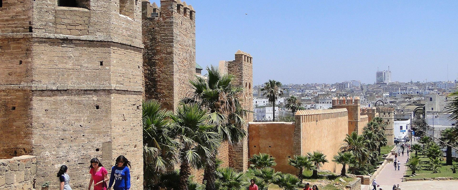 A view of ancient stone fortifications with crenellated towers and a wide paved walkway lined with palm trees, with pedestrians strolling along the path and a modern city skyline visible in the background.