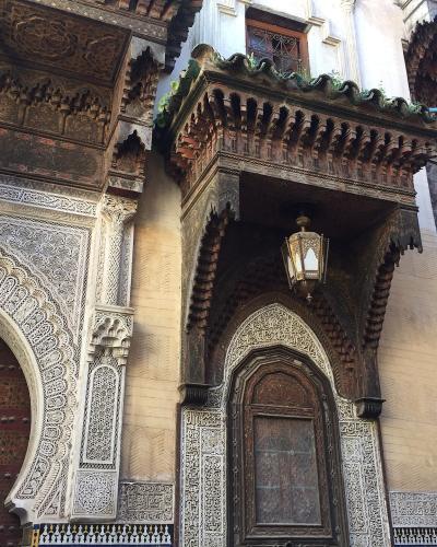 The image shows the ornate facade of a historic religious building in Morocco, featuring intricate zellige tilework, carved stucco, and a projecting wooden balcony (mashrabiya) above an arched doorway.