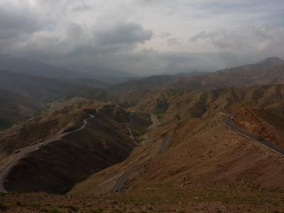A winding mountain road snakes through arid, rolling hills under a cloudy sky, with vehicles visible on the asphalt.