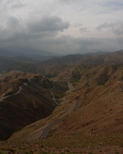 A winding mountain road snakes through arid, rolling hills under a cloudy sky, with vehicles visible on the asphalt.