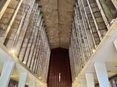 A low-angle interior shot of a modernist church nave featuring tall, ribbed concrete walls and a vaulted ceiling, with a crucifix hanging centrally above a wooden altar area.