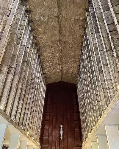 A low-angle interior shot of a modernist church nave featuring tall, ribbed concrete walls and a vaulted ceiling, with a crucifix hanging centrally above a wooden altar area.