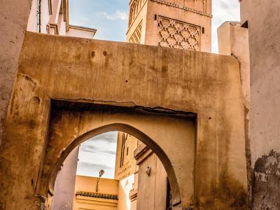 A low-angle view of a weathered stone archway in the foreground framing a tall, square brick minaret with geometric zellige tilework and a finial, set against a blue sky with clouds.
