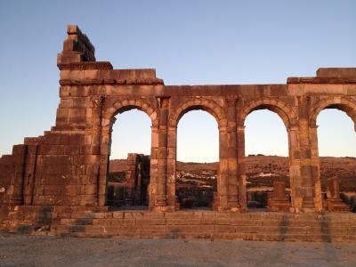 The image shows the stone facade of an ancient Roman triumphal arch with four arches, set against a clear sky with hills in the background.