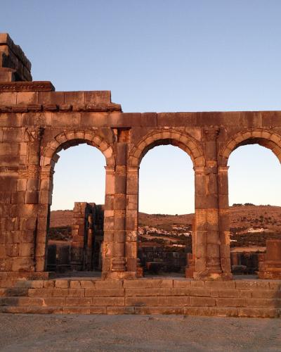 The image shows the stone facade of an ancient Roman triumphal arch with four arches, set against a clear sky with hills in the background.