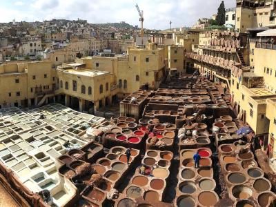 A high-angle panoramic view of a traditional leather tannery featuring rows of circular stone vats filled with colored liquids and workers processing hides, surrounded by yellow-walled buildings in a dense urban medina setting.