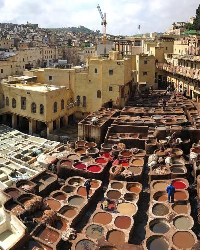 A high-angle panoramic view of a traditional leather tannery featuring rows of circular stone vats filled with colored liquids and workers processing hides, surrounded by yellow-walled buildings in a dense urban medina setting.