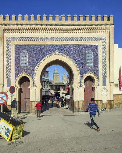 A large, ornate city gate with blue and white zellige tilework, a central horseshoe arch, and crenellated parapet, with people walking through and around it under a clear blue sky.