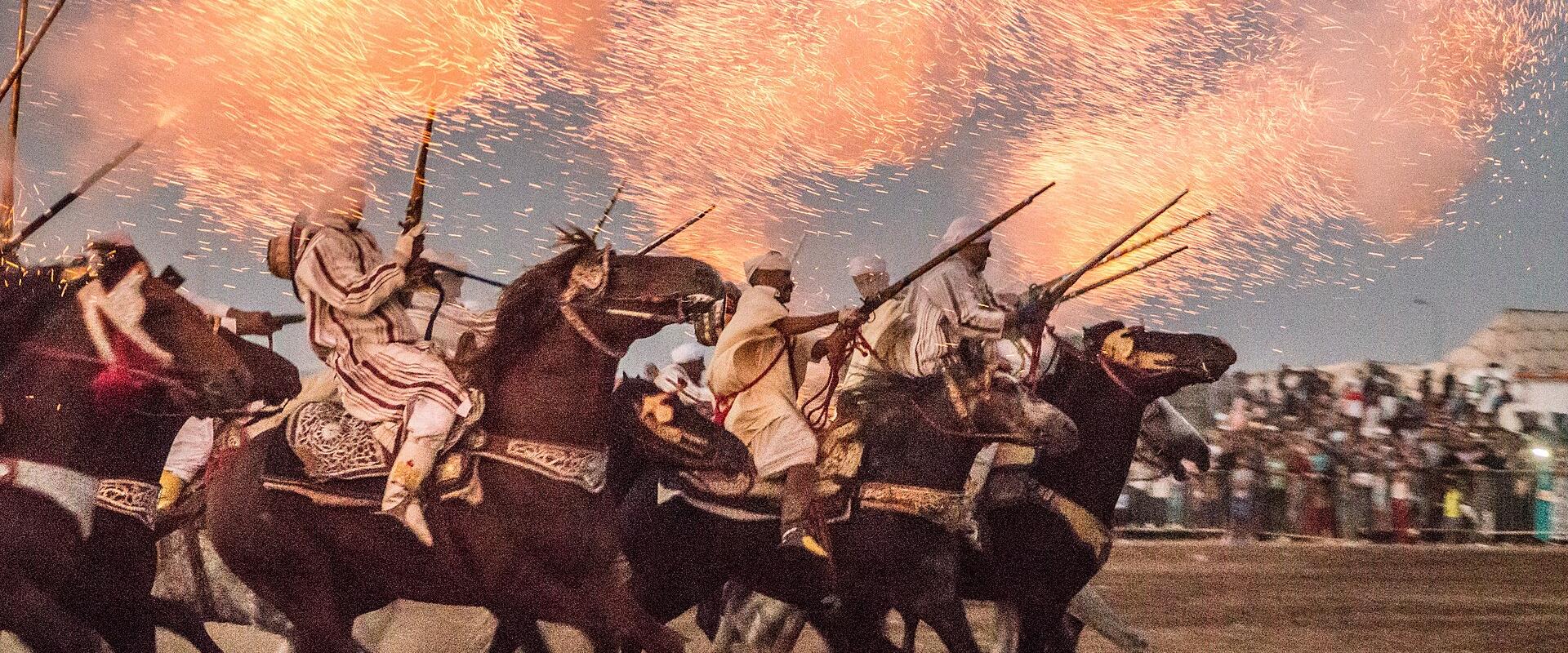 A group of riders on horseback performing a traditional Moroccan equestrian display (Tbourida) with fireworks or sparks erupting above them in an outdoor arena.