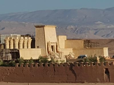 A wide shot of a large, fortified film set entrance with Egyptian-style statues and an arched gateway, set in a desert landscape with mountains in the background.