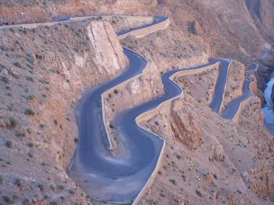 A high-angle view of a winding paved road with hairpin turns carved into a steep, arid mountainside, with a river visible in the gorge below.