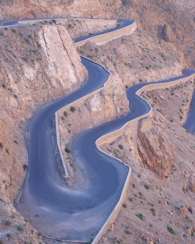 A high-angle view of a winding paved road with hairpin turns carved into a steep, arid mountainside, with a river visible in the gorge below.
