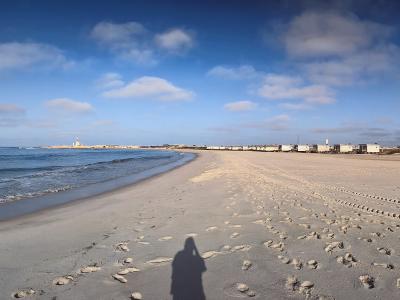 A panoramic view of a wide sandy beach along the Atlantic coast, featuring a long breakwater with a lighthouse in the distance and a row of low-rise buildings along the shoreline.