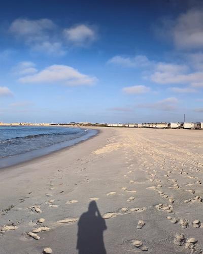 A panoramic view of a wide sandy beach along the Atlantic coast, featuring a long breakwater with a lighthouse in the distance and a row of low-rise buildings along the shoreline.