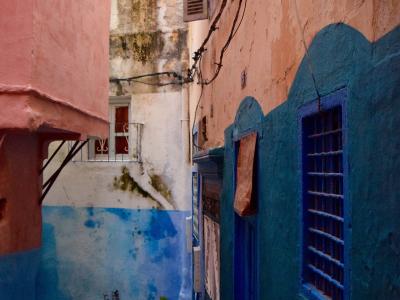 A narrow alleyway between buildings with pink and blue painted walls, featuring a blue wooden door and window with bars, and a street lamp overhead.