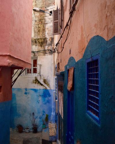 A narrow alleyway between buildings with pink and blue painted walls, featuring a blue wooden door and window with bars, and a street lamp overhead.