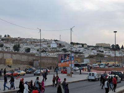 A busy street scene in front of a high city wall, with a large cemetery of white tombs covering the hillside behind the wall and pedestrians crossing the road.