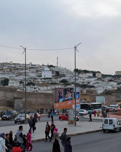 A busy street scene in front of a high city wall, with a large cemetery of white tombs covering the hillside behind the wall and pedestrians crossing the road.