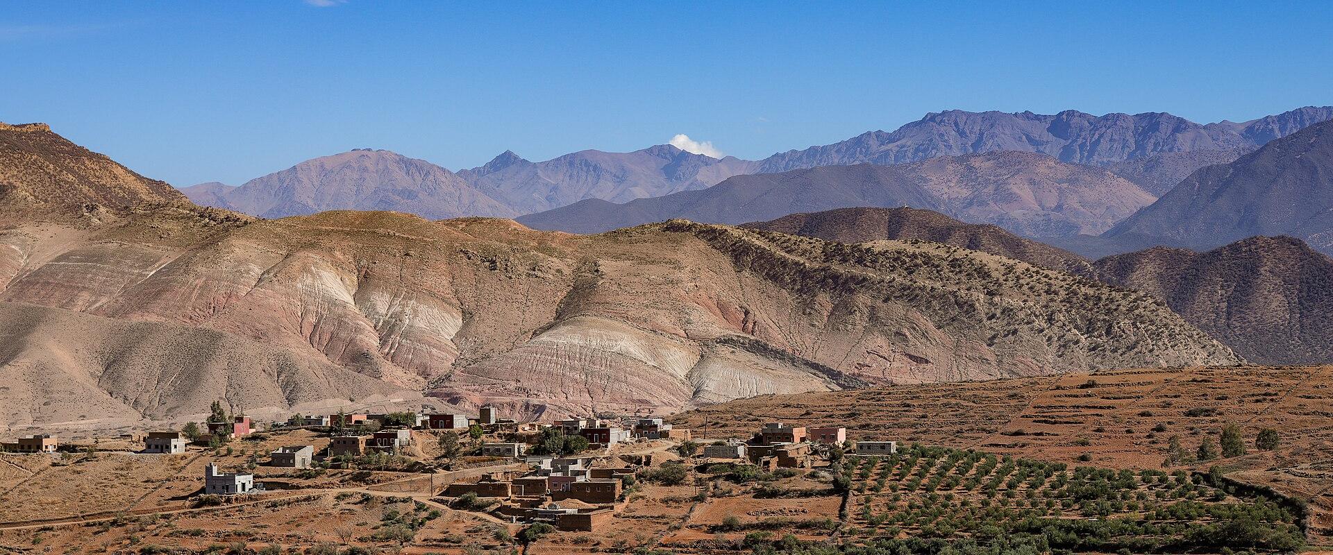 A rural village with traditional earthen buildings nestled in a valley, surrounded by arid, rolling hills and layered mountain ranges under a blue sky.