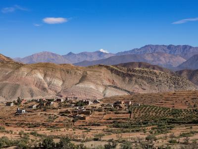 A rural village with traditional earthen buildings nestled in a valley, surrounded by arid, rolling hills and layered mountain ranges under a blue sky.