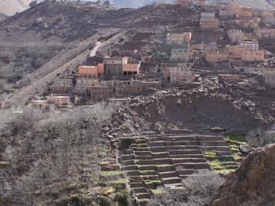 A village of stone and mud-brick houses built on a steep, rocky mountainside with terraced agricultural fields in the foreground and snow-capped peaks in the background.