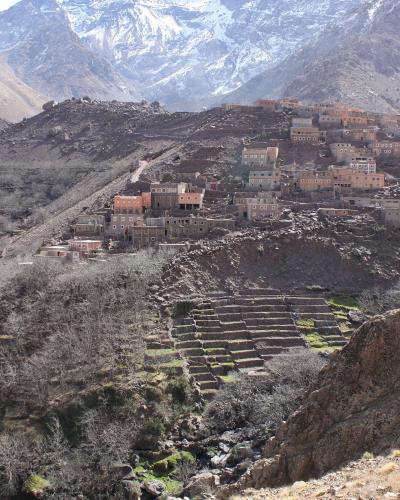 A village of stone and mud-brick houses built on a steep, rocky mountainside with terraced agricultural fields in the foreground and snow-capped peaks in the background.