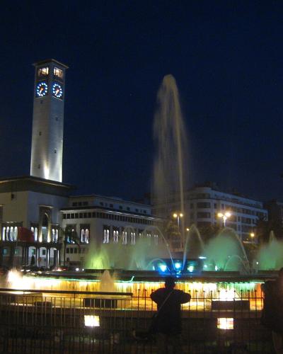 A night view of a large public square featuring a prominent clock tower building on the left and a large illuminated fountain in the center foreground.