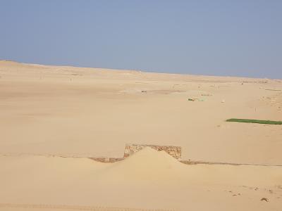 A wide shot of a sandy desert landscape featuring a small stone structure in the foreground and several green artificial turf mats or targets set up in the sand dunes under a clear blue sky.