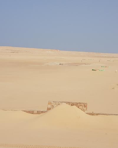 A wide shot of a sandy desert landscape featuring a small stone structure in the foreground and several green artificial turf mats or targets set up in the sand dunes under a clear blue sky.
