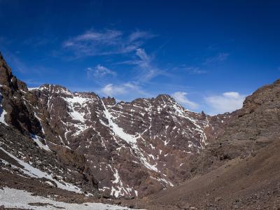 A high-altitude mountain valley with steep rocky slopes and patches of snow under a blue sky.
